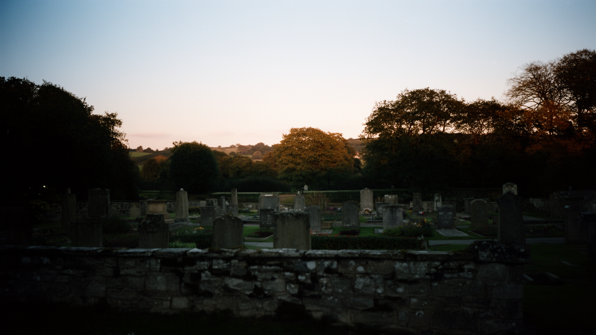 A quiet Yorkshire memorial garden at dusk, weathered stone in the foreground and a calm tree-lined horizon